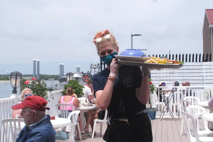 A masked waitress carries a tray on her shoulder and walks between tables of massless outdoor diners with the ocean in the background